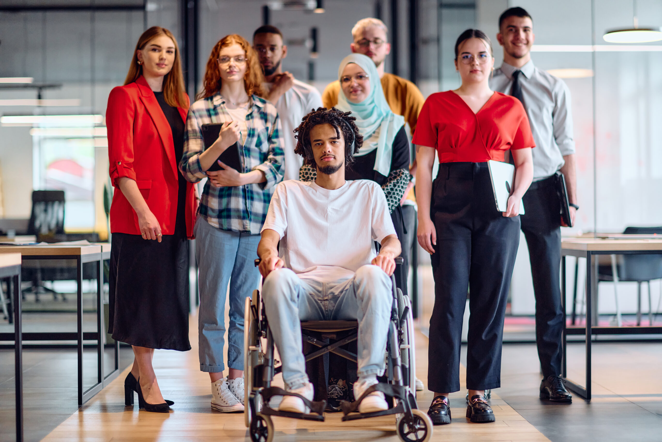 A diverse group of young business people walking a corridor in the glass-enclosed office of a modern startup, including a person in a wheelchair and a woman wearing a hijab