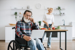 Old man sits in wheelchair and works on laptop while his wife prepares healthy salad.